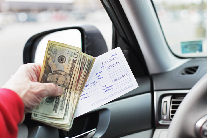 A man in his car is holding five hundred dollars in US twenties and a bank deposit slip while waiting in line for a bank drive through teller window